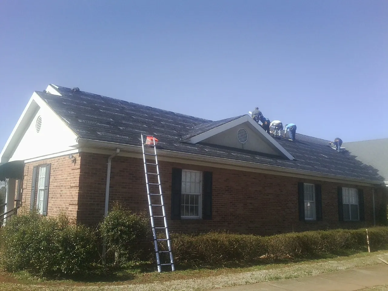 Crew tearing shingles off a brick ranch roof with a ladder set — Metal Roof Repair work by our crew in Cedar Valley