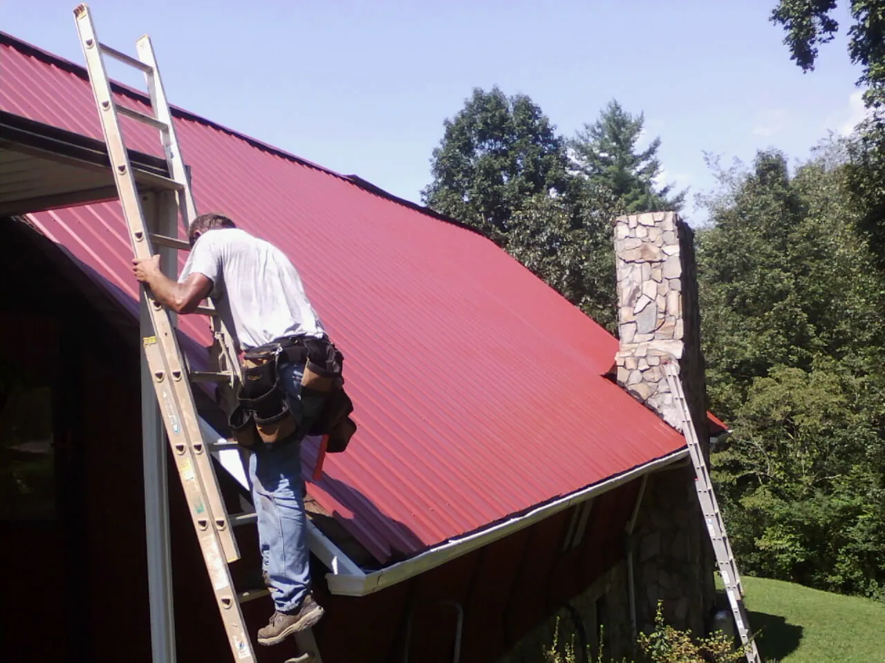 Roofer on a ladder beside a red metal roof and stone chimney — Soffit & Fascia Repair by Roofing Brothers in Cedar Valley